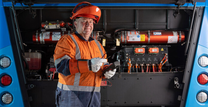 Man in high-visibility overalls and headwear stands in front of electric bus