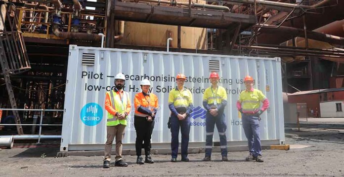 Men in suits and high-vis vests and safety gear pose for photo in front of shipping container at steelworks plant