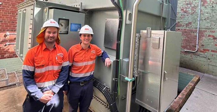 Two male Essential Energy employees wearing high-vis safety gear and hard hats stand next to a transformer