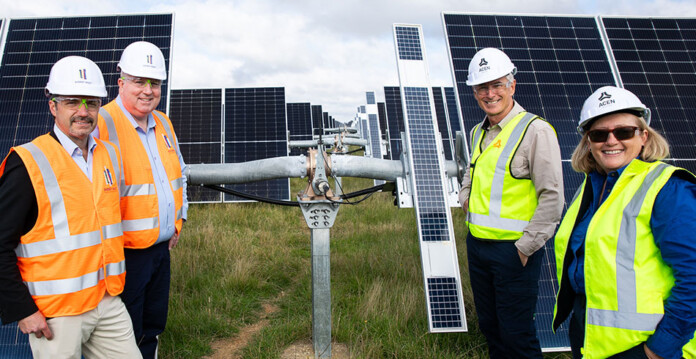 Executives in high-vis vests and hard hats stand in front of solar panels at New England Solar Farm