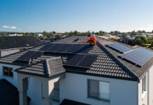 Ombudsman says solar and battery complaints have doubled Solar installer in safety gear on roof of double-storey house with suburb in background