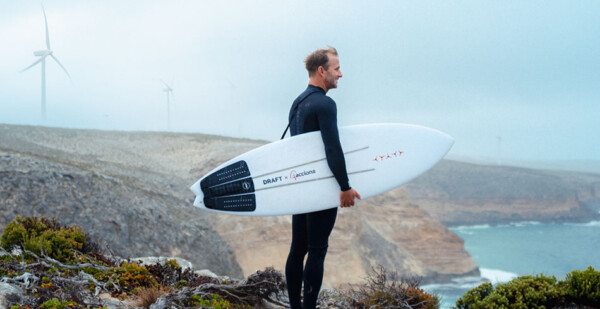 Male surfer stands with surfboard on beachside cliff with wind turbines in background