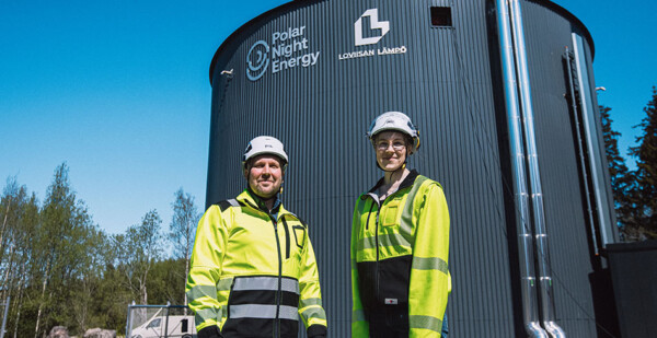 Two Polar Night Energy employees in safety gear stand in front of massive Sand Battery with blue sky and trees in the background