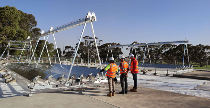 Three people in safety vests and hard hats stand in front of photocatalytic water splitting equipment with blue sky and trees in background