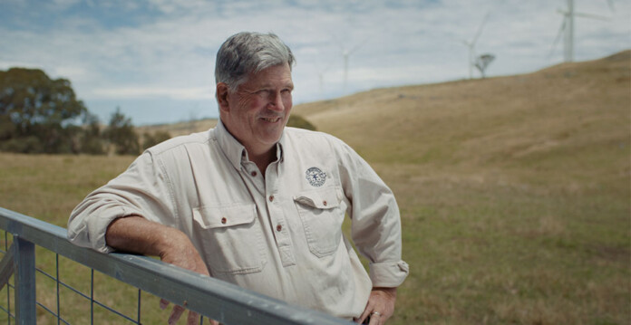 Smiling grey-haired farmer leans on fence with paddock and wind turbines in background