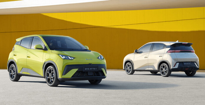 Two BYD Atto 1 models, one lime-yellow and one white, in front of orange wall