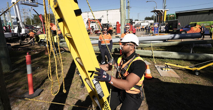 Powerline worker in hard hat and safety harness climbs large yellow ladder