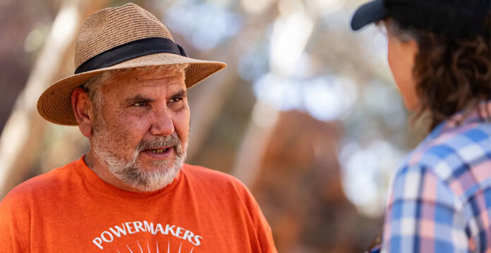 First Nations man in hat and orange shirt bearing PowerMakers logo chats to another man in a checked shirt