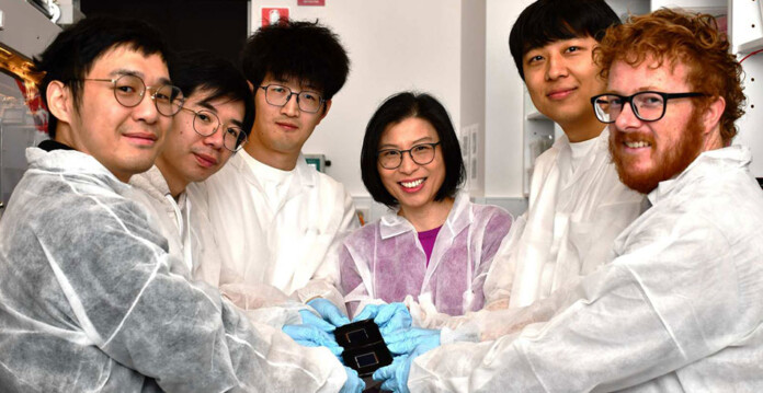 Group of people in lab coats wearing gloves holding tiny solar cell smiling for photo