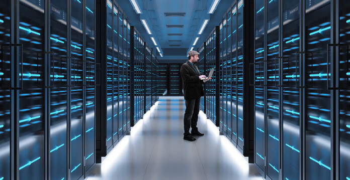 A man stands in the aisle of a data centre lit with blue lights