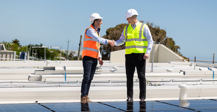 Two men in hard hats and high-vis vests over businesswear shake hands on top of roof with solar panels