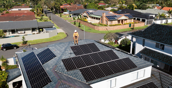 Solar installer wearing safety gear and hardhat stands on top of roof with solar panels in suburban street