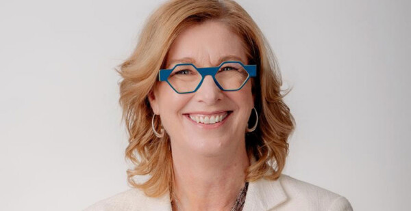 Head and shoulders shot of smiling woman with blonde hair and contemporary glasses wearing a white jacket