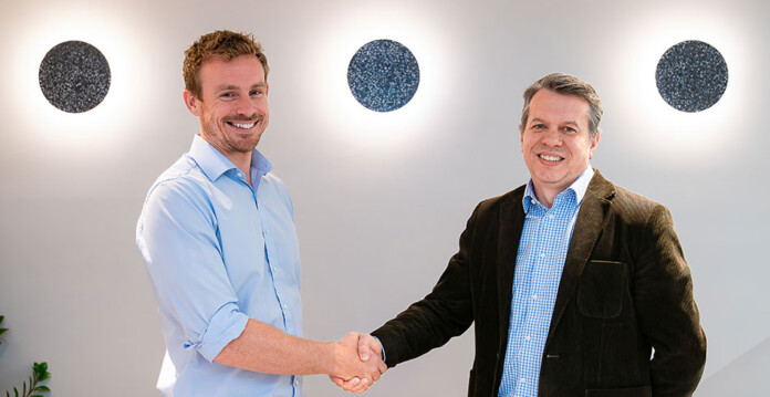 Two men pose for photo while shaking hands in brightly lit office