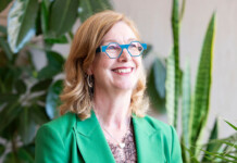 Spotlight on: AEMC chair Anna Collyer Close-up of blonde woman wearing blue framed glasses and vibrant green suit with plants in background