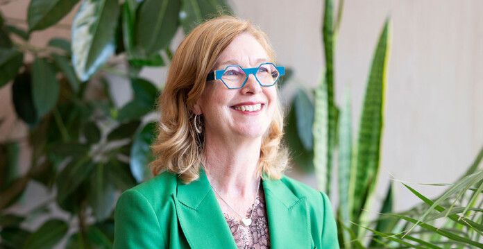 Close-up of blonde woman wearing blue framed glasses and vibrant green suit with plants in background