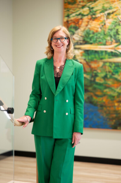 Photo of woman in stairwell wearing blue framed glasses and vibrant green suit, with a large painting in the background