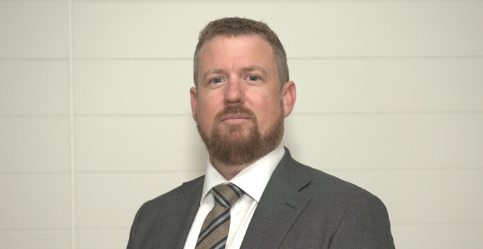 greg-elkins Head and shoulders shot of bearded man wearing a tie and dark grey suit