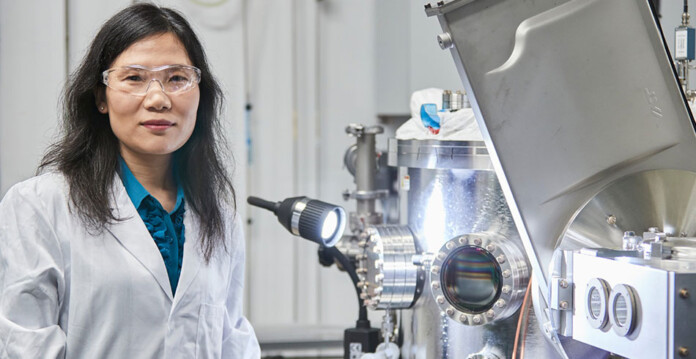 Female researcher in lab coat and safety goggles stands next to microscope in lab