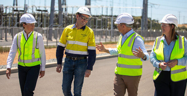 Executives in high-vis vests and white hard hats walk through large battery energy storage system