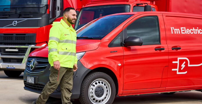 Man in high-vis yellow jacket walks in front of electric Australia Post vehicles