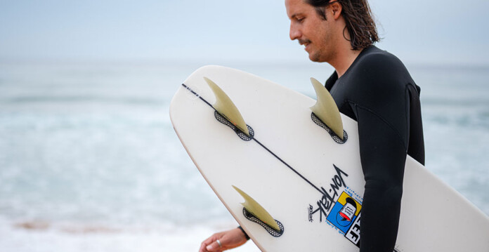 Young male surfer in black wetsuit holds surfboard while walking into surf