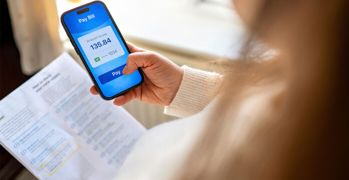 Woman with long brown hair holds energy bill and smartphone with bill payment amount on screen