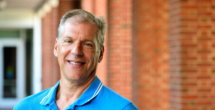 Head and shoulders shot of smiling man wearing blue shirt standing in front of brick building