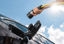 Why consumers and cars are key to energy independence Upward view of female hand holding EV charger next to vehicle with blue sky and sun shining overhead