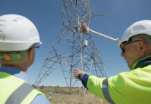 Transmission towers to fix mobile coverage gaps in regions Three energy workers in high-vis vests and white hardhats gesture towards construction of a giant transmission tower in a rural landscape setting