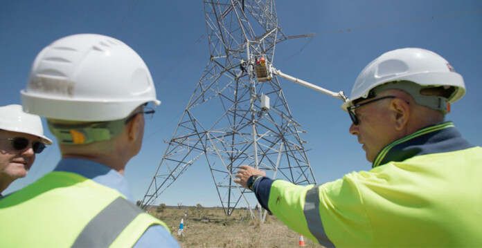 Three energy workers in high-vis vests and white hardhats gesture towards construction of a giant transmission tower in a rural landscape setting