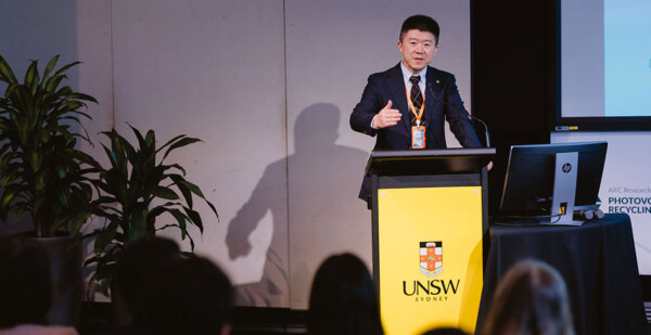 Man in black suit stands at lectern with UNSW logo in front of audience