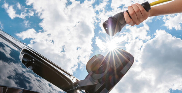 Hand holding EV charger up to electric vehicle with blue sky and clouds overhead