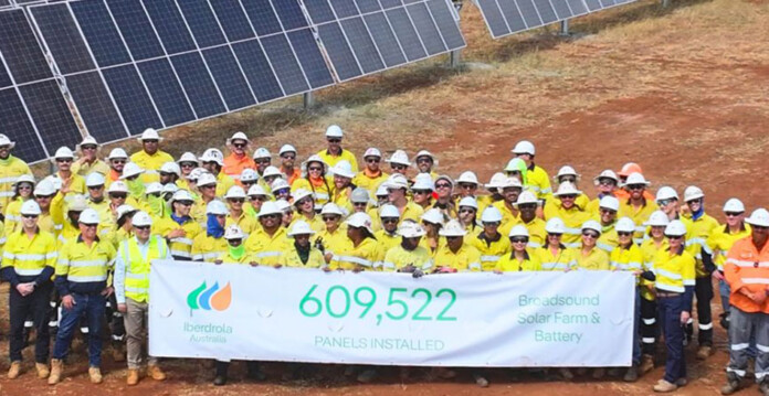 Huge construction crew in hard hats and high-vis vests stand in front of solar panels at solar farm holding sign announcing completion of solar installation at Broadsound Solar Farm