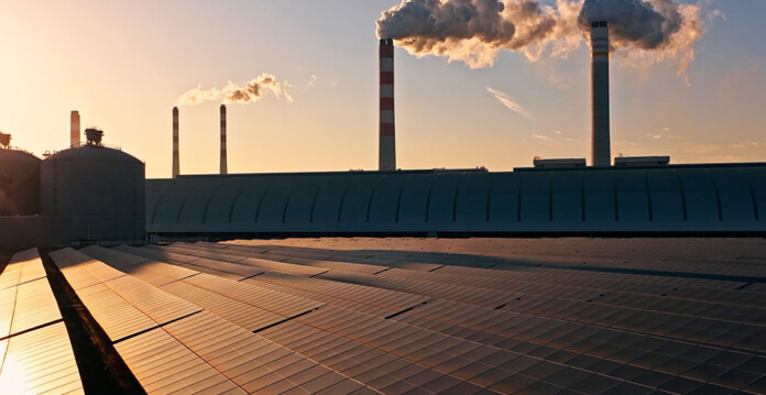 Solar panels in front of coal-fired power plant with smoke pouring from chimneys