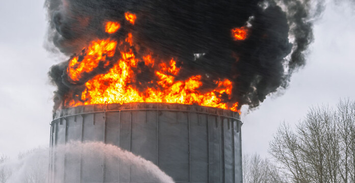 Flames and black smoke pour from oil refinery storage tank, with stream of water underneath