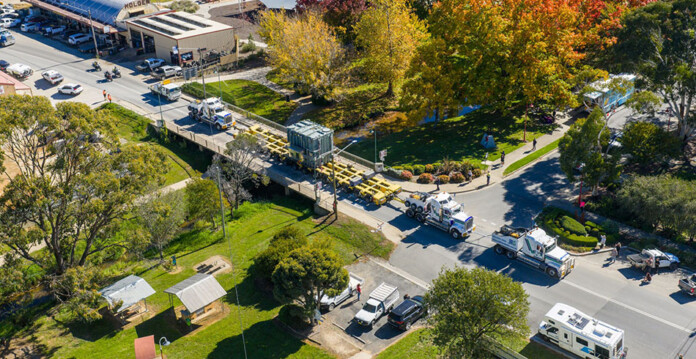 Aerial shot of a convoy transporting large pieces of transmission equipment along a road with trees and buildings on either side