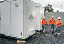 Ausgrid launches new community battery on Central Coast three energy workers in hard hats and high-vis vests walk beside large white community battery in fenced-off area
