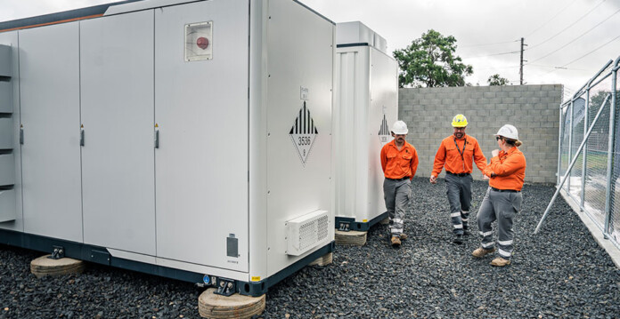 three energy workers in hard hats and high-vis vests walk beside large white community battery in fenced-off area