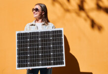‘Right to plug in’ proposals target cheaper power for renters Smiling woman wearing sunglasses holds portable solar panel in front of sunny orange wall