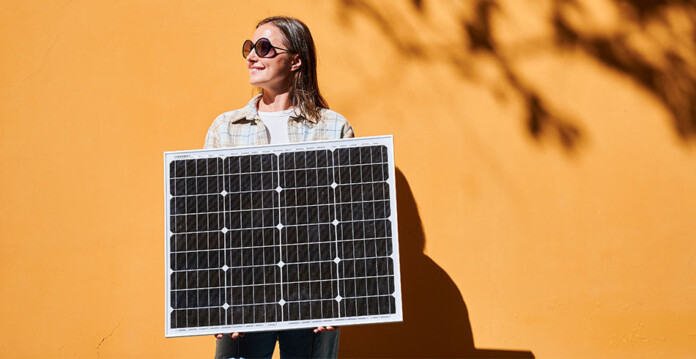 Smiling woman wearing sunglasses holds portable solar panel in front of sunny orange wall
