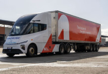 New Energy Transport accelerates electric road freight fleet Massive red and white electric prime mover truck with warehouse behind it