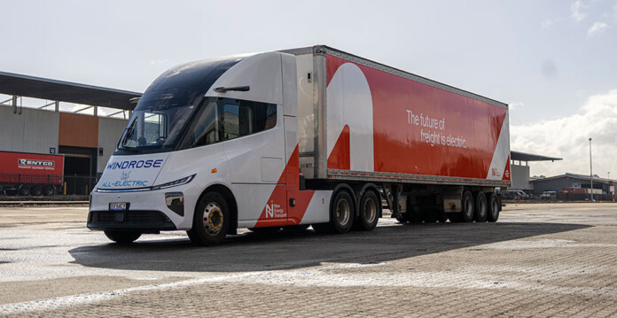 Massive red and white electric prime mover truck with warehouse behind it