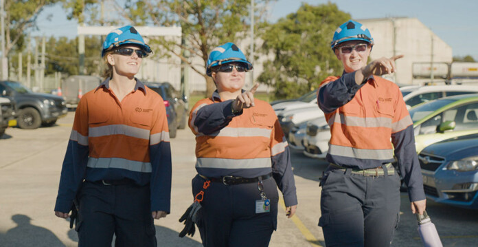 Three young female energy workers walking together outside wearing caps and high-vis workwear
