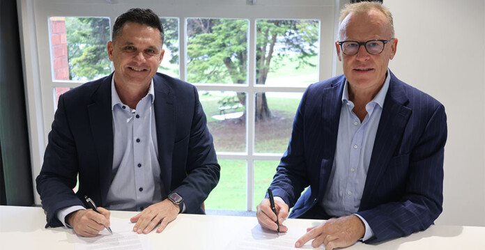 Two male executives sign agreement papers at a desk in a sunlit room