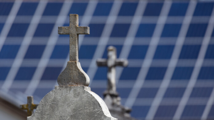 Crosses on the headstone of graves in the foreground with distant solar panels blurred in the background