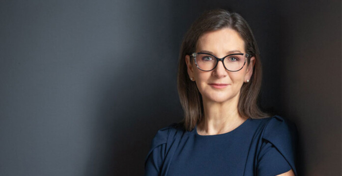 Head and shoulders shot of a woman with dark brown hair and dark-framed eyeglasses in a navy blouse leaning against a dark wall