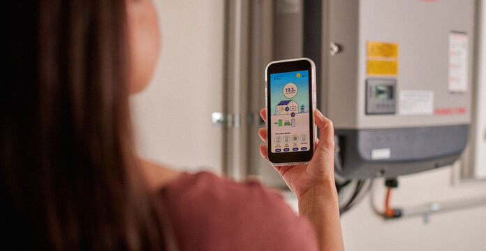 Woman with long dark hair holds smart phone with energy usage app while standing in front of solar inverter