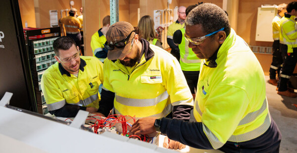 Three men in electrical safety wear stand at a bench where an electrical apprentice works on a project