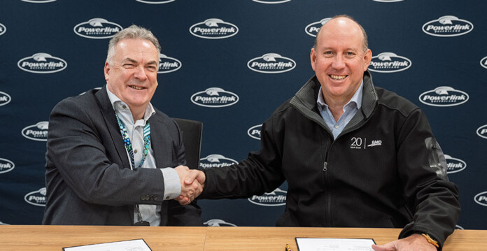 Two men sit in front of wall with Powerlink logo and shake hands for photo